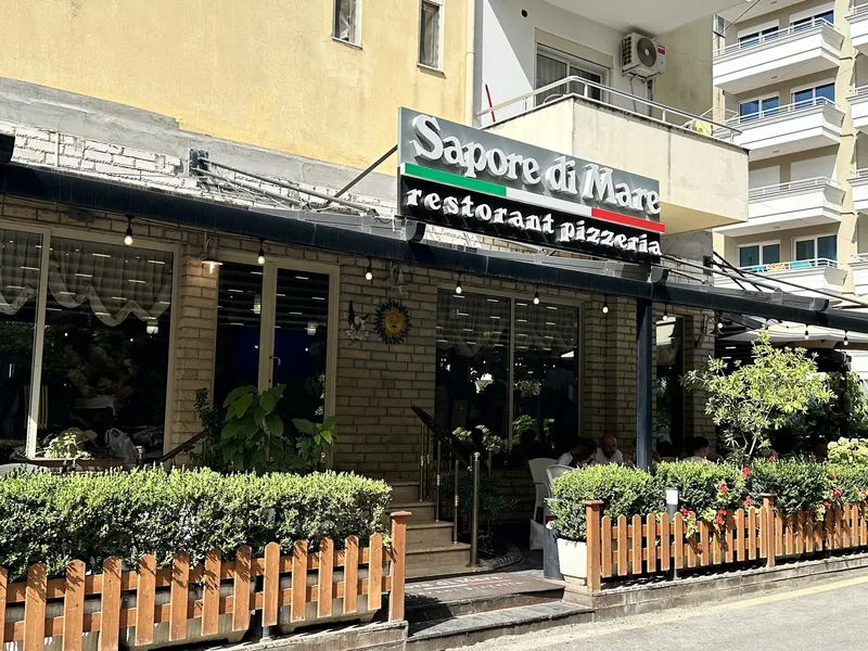 The front of Sapore Di Mare in daylight, with its sign above a terrace framed by a hedge and a wooden fence